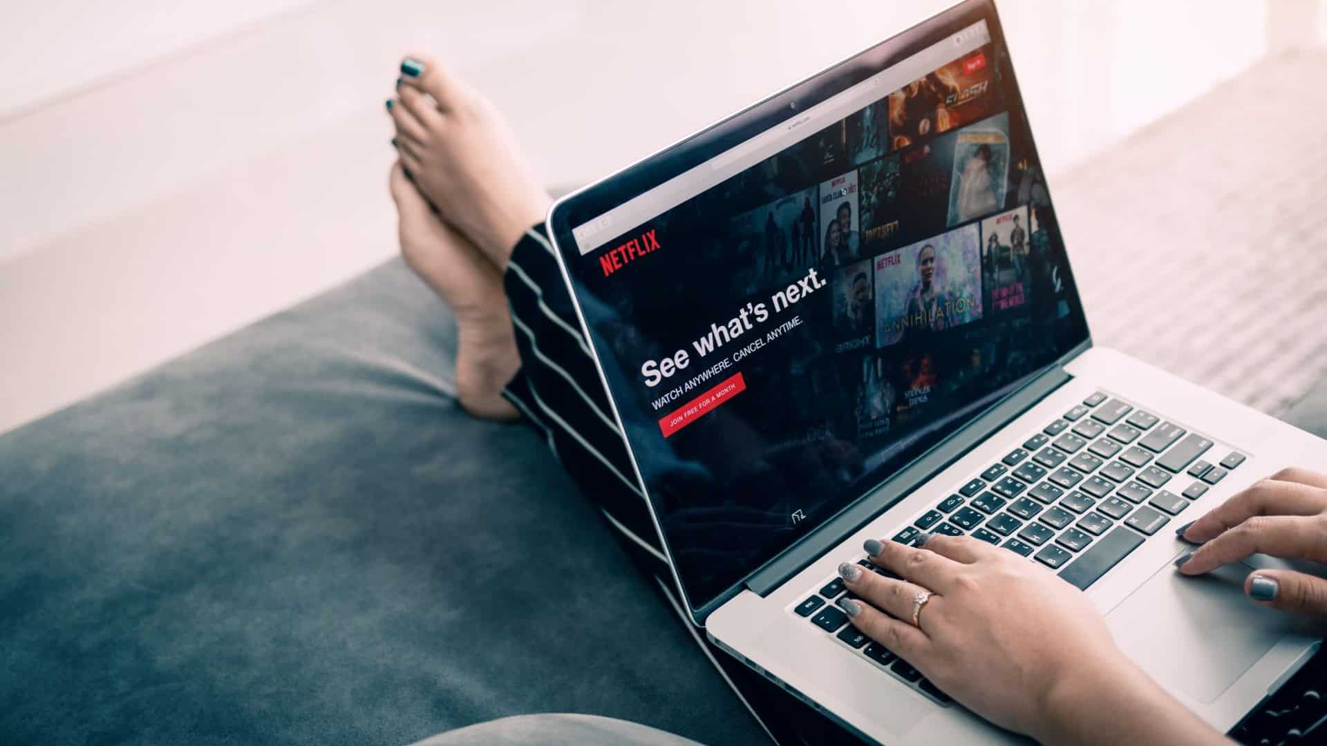 Pessoa sentada no sofá com pés estendidos e notebook no colo, onde acessa a Netflix