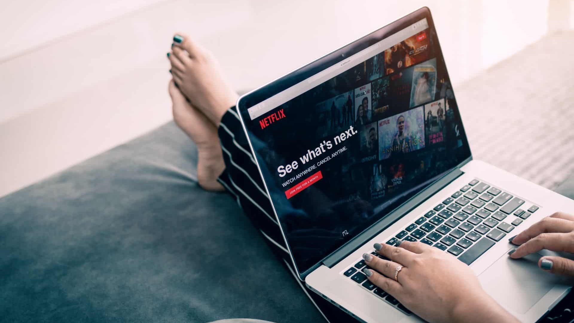 Pessoa sentada no sofá com pés estendidos e notebook no colo, onde acessa a Netflix