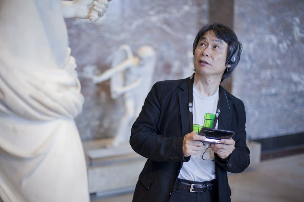 imagem do shigeru miyamoto, criador do super mario, olhando para uma estatua com um videogame nas mãos