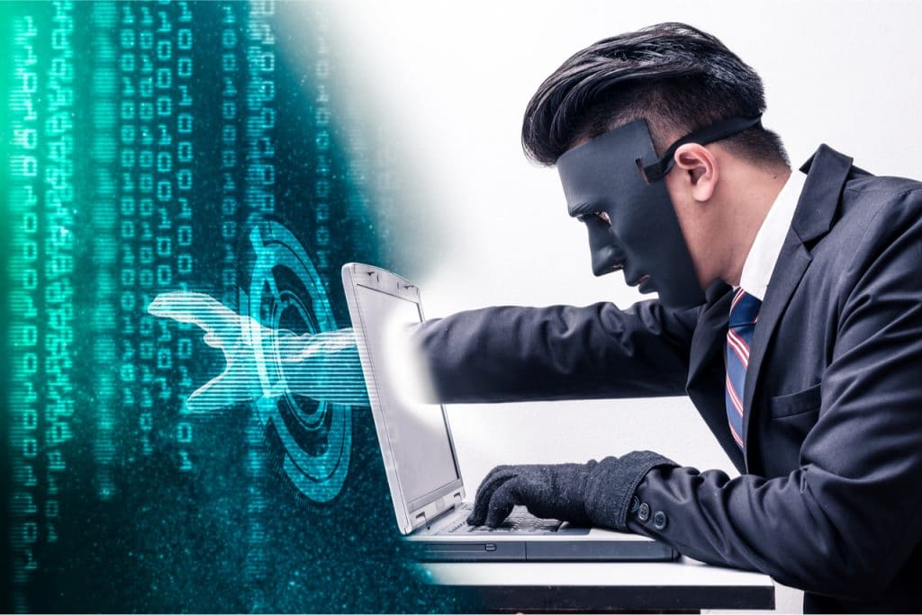 Imagem conceitual mostra um hacker mascarado esticando a mão dentro de um computador, para roubar uma base de dados sigilosos