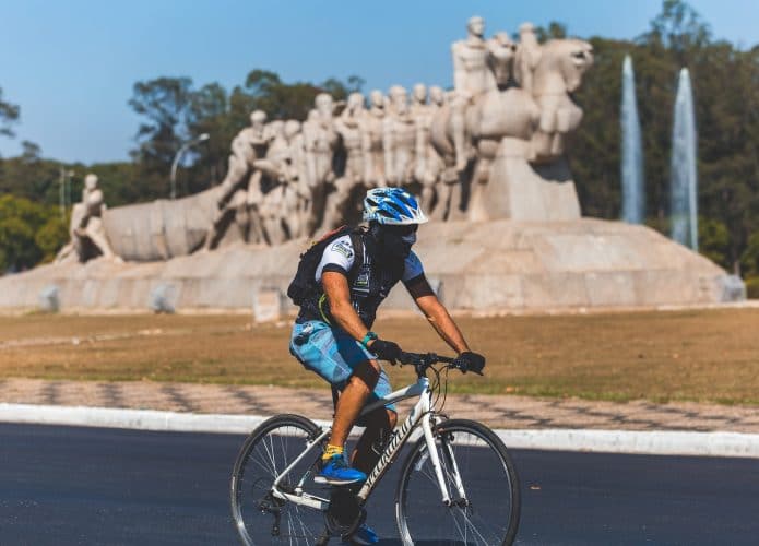Homem anda de bicicleta com máscara facial nas ruas desertas de São Paulo durante surto de Coronavírus e quarentena com Monumento as Bandeiras ao fundo