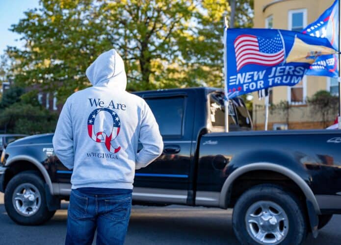 Homem encapuzado vestindo um moletom Qanon em uum comício de Donald Trump com um caminhão e bandeiras à sua frente