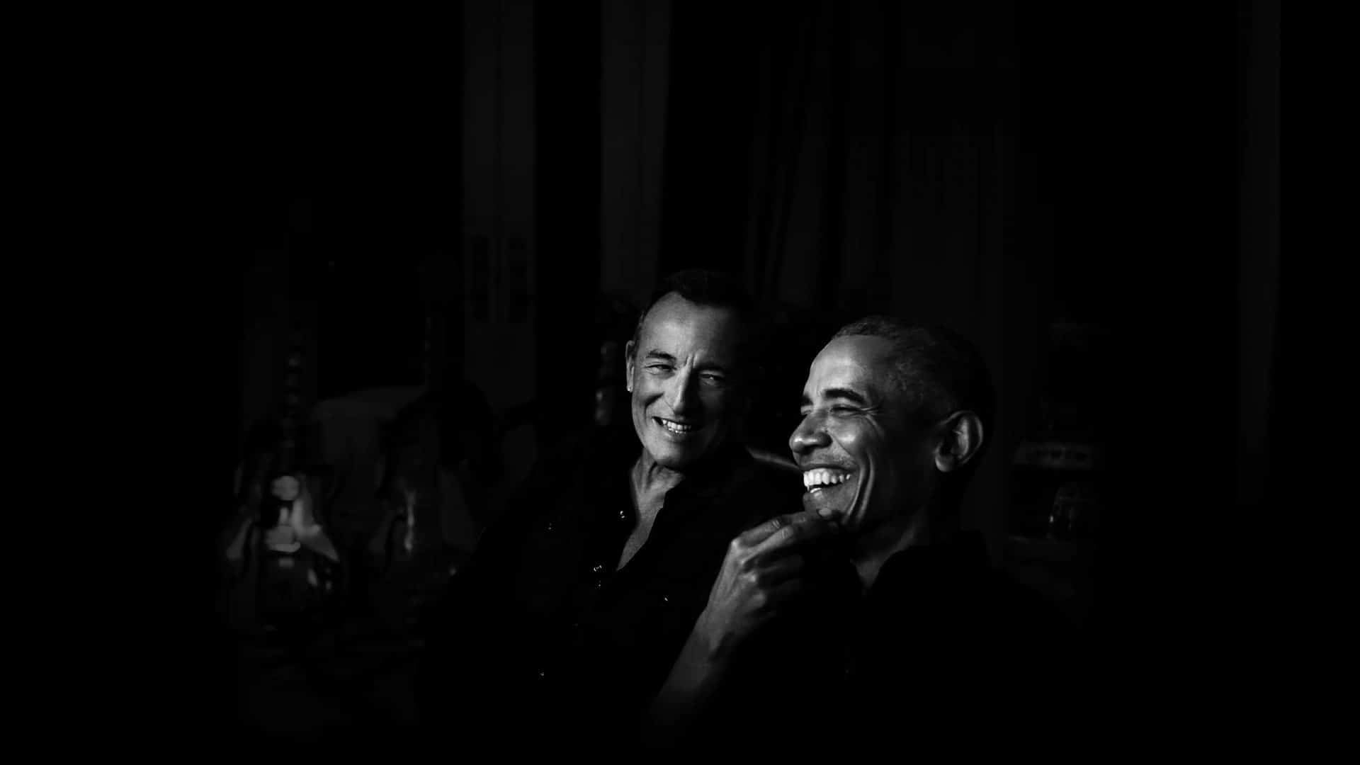Barack Obama e Bruce Springsteen aparecem sorrindo, em uma foto preto-e-branco com o fundo bem escuro