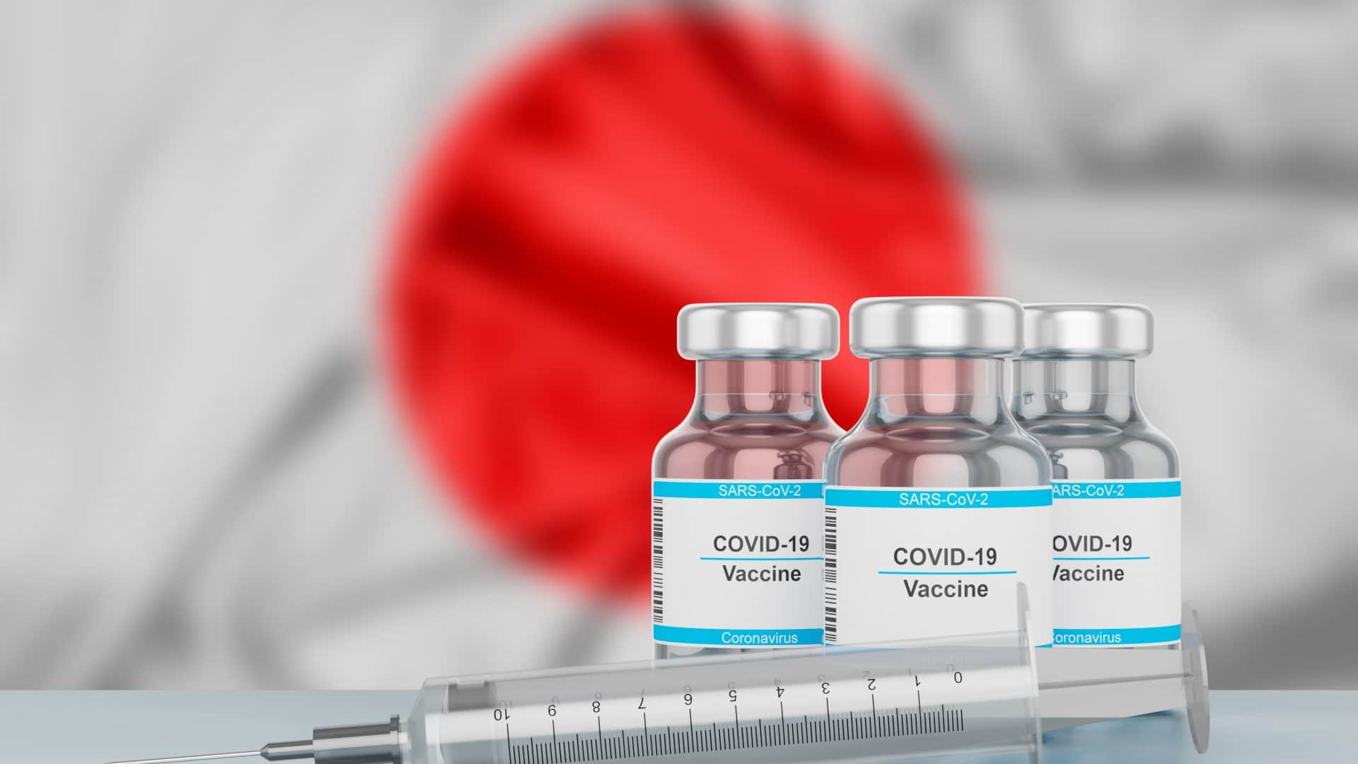 Bandeira do Japão desfocada ao fundo, frascos de vacina e seringa em primeiro plano.