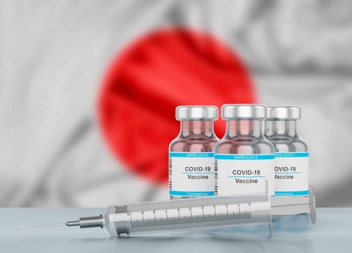 Bandeira do Japão desfocada ao fundo, frascos de vacina e seringa em primeiro plano.