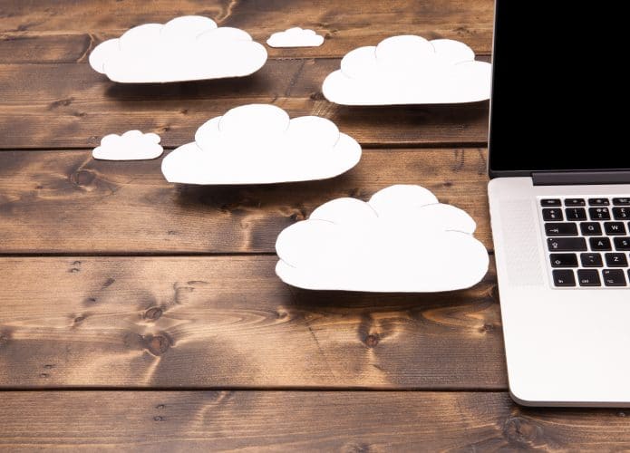 Imagem mostra uma parte de um notebook, em cima de uma superfície de madeira, e ao lado dele algumas nuvens feitas de papel. A ideia é ilustrar a computação em nuvem.