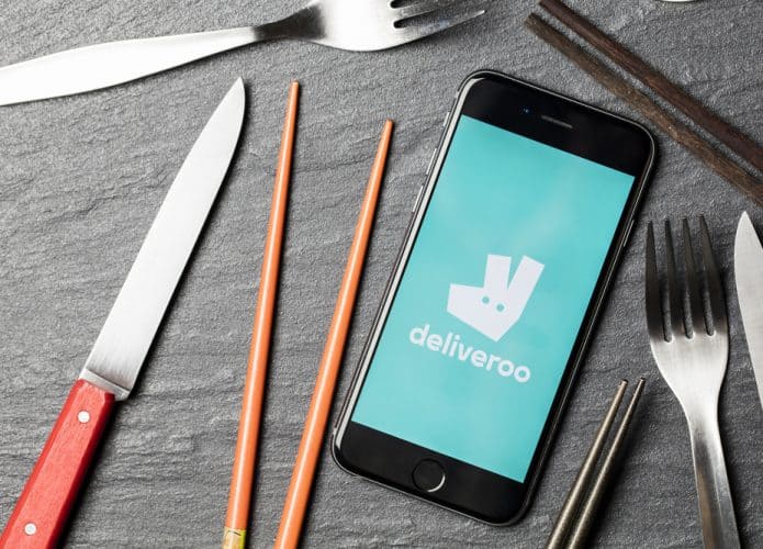 Sobre uma mesa está um smartphone com o logo do aplicativo de delivery Deliveroo e, ao lado, talheres e hashis