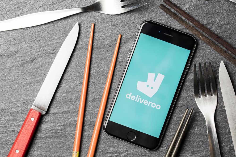 Sobre uma mesa está um smartphone com o logo do aplicativo de delivery Deliveroo e, ao lado, talheres e hashis