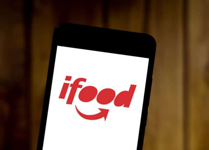 Smartphone mostra logo do iFood na tela. A foto foi tirada em um local com fundo de madeira.