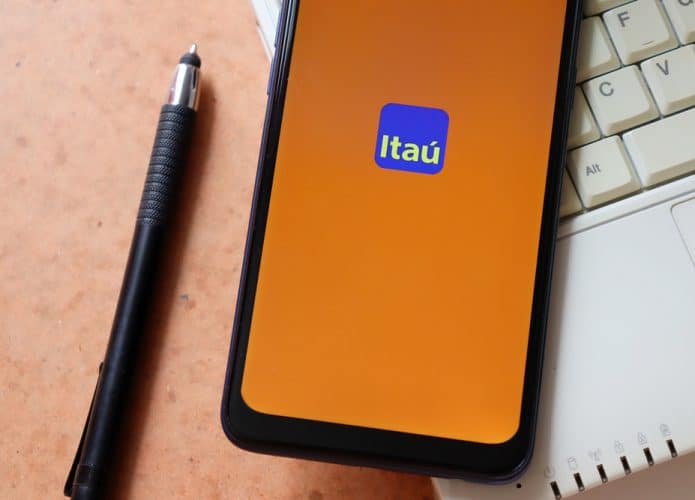 Logotipo do banco Itaú aparece em tela de smartphone, que por sua vez está em cima de um teclado de notebook e ao lado de uma caneta.