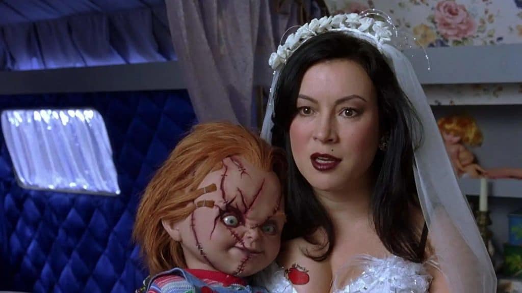 Imagem mostra a atriz Jennifer Tilly, que participou do filme "A Noiva de Chucky". Ela está vestida de noiva, com maquiagem gótica e cabelo preto, carregando o boneco titular em seu colo