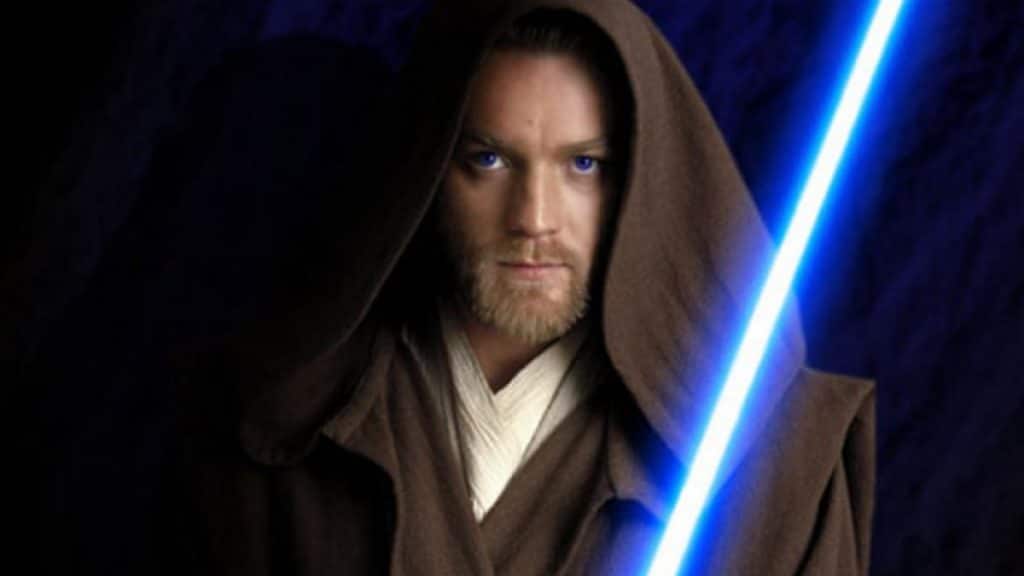 Image mostra Ewan McGregor interpretando Obi-Wan Kenobi: personagem está vestindo um capuz marrom escuro e segurando um sabre de luz de cor azul