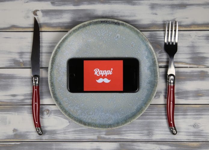 A imagem mostra um prato acompanhado com talheres sobre uma mesa. No prato, um celular com o logotipo do Rappi na tela