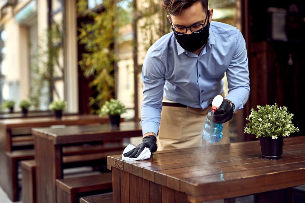 Garçom realiza limpeza das mesas de um restaurante durante a pandemia, utilizando máscara e luvas.