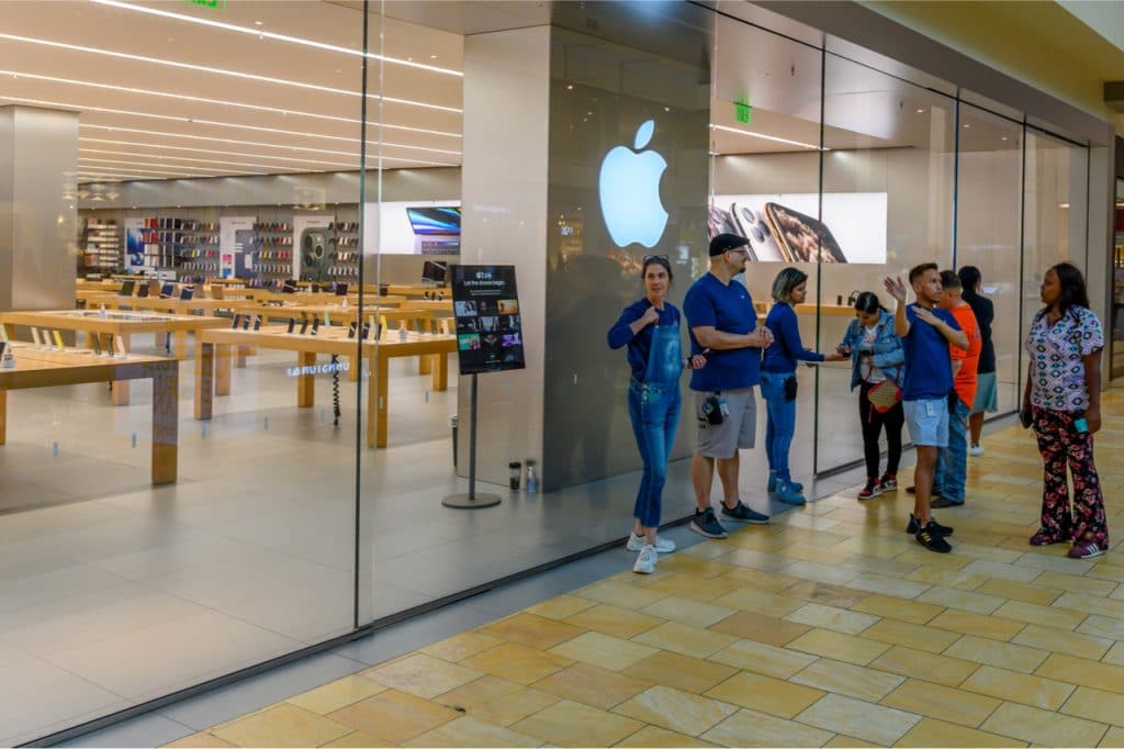 Imagem mostra a logomarca da Apple na fachada de uma loja da empresa, com funcionários à frente medindo temperaturas de consumidores antes que estes entrem no local