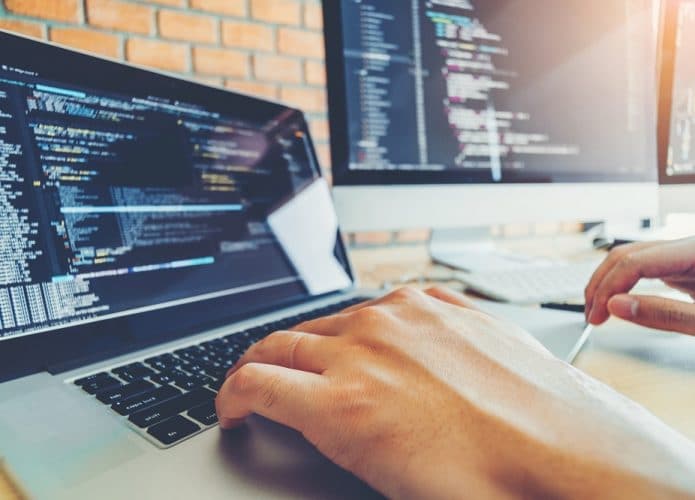 Imagem mostra uma mão digitando um código de programação em um teclado de notebook, ao lado a tela de um iMac.