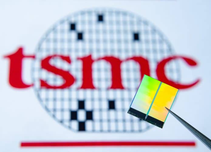 Imagem mostra ao fundo o logotipo da Fabricante de chips TSMC, à frente há uma pinça segurança um chip