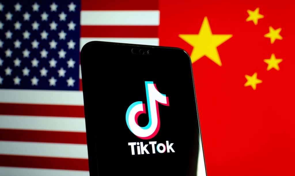 Bandeiras dos EUA e China ao fundo de um smartphone exibindo o logo do TikTok