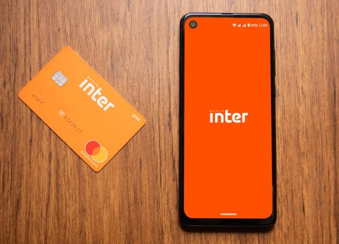 Em uma superfície de madeira, há um cartão do Banco Inter e, ao lado, um smartphone exibindo o logo do mesmo banco na tela.