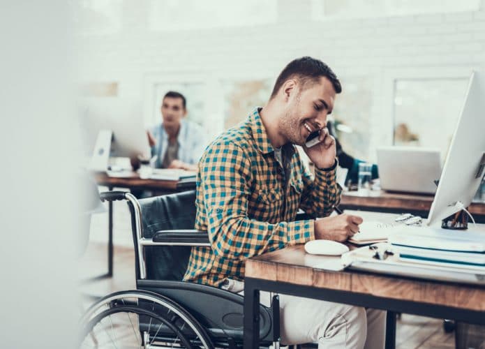 PCD na cadeira de rodas está alocado em uma mesa, mexendo no computador e falando ao telefone