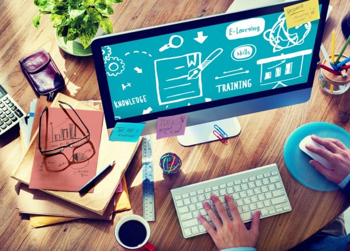 Imagem ilustra treinamento e capacitação em tecnologia, com uma mesa cheia de papeis e um monitor acompanhado de mouse e teclado.
