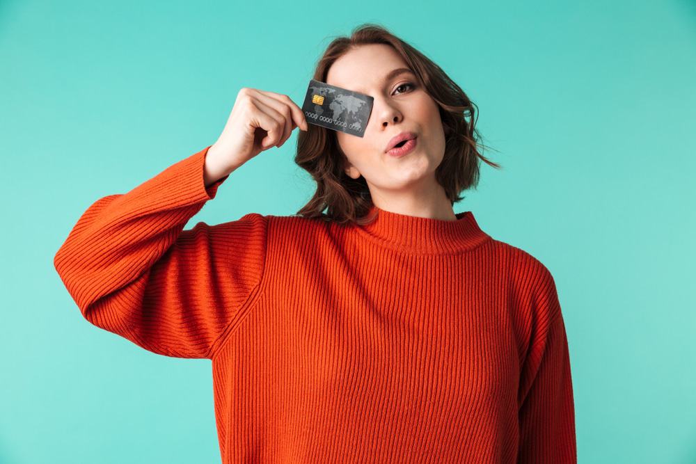 A imagem com um fundo verde claro mostra uma jovem de blusa laranja, segurando um cartão de crédito na mão, tapando um dos olhos.