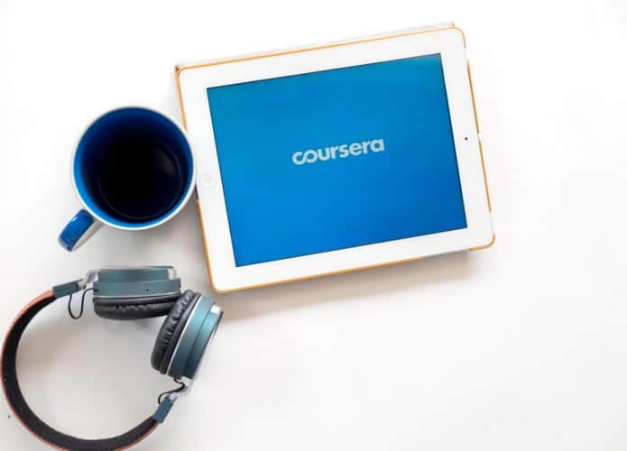 Em uma superfície estão um fone de ouvido headset, uma canela e um tablet com o logo da edtech Coursera na tela.