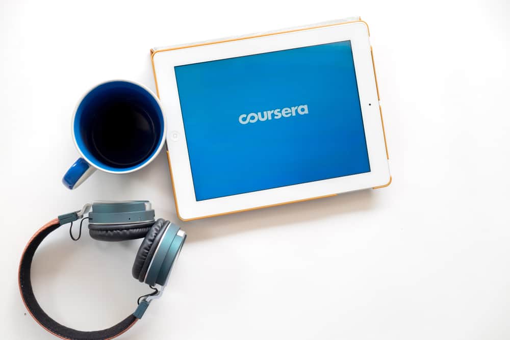 Em uma superfície estão um fone de ouvido headset, uma canela e um tablet com o logo da edtech Coursera na tela.