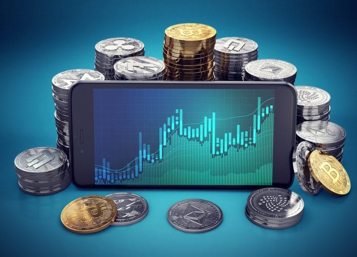 Imagem exibe um celular com a tela cheia de gráficos; ao fundo, pilhas de diferentes criptomoedas