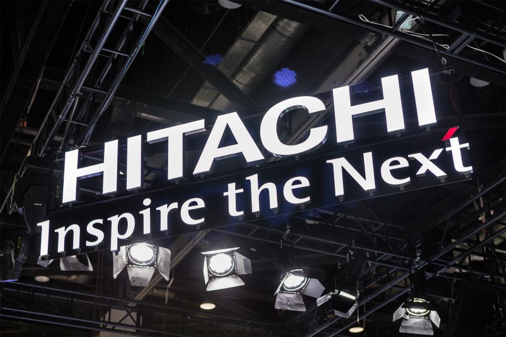 Logo da Hitachi em letra caixa, pendurado em um teto; abaixo o slogan Inspire the Next