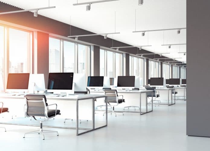 Imagem mostra uma sala aberta de escritório, com janelas grandes à esquerda, mesas enfileiradas e acompanhadas de cadeiras de escritório giratórias, além de PCs e notebooks em cima das mesas.