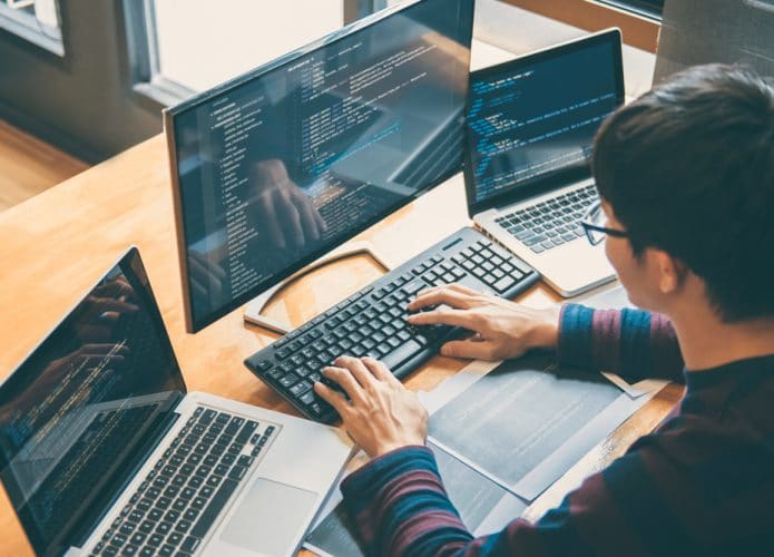 Homem sentado em uma mesa com três notebooks, programando