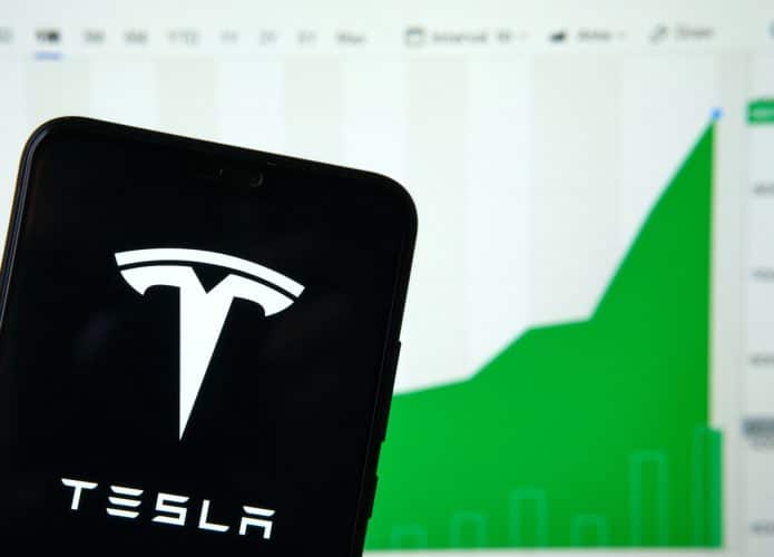 Logo da Tesla em um smartphone e ao fundo as ações da empresa