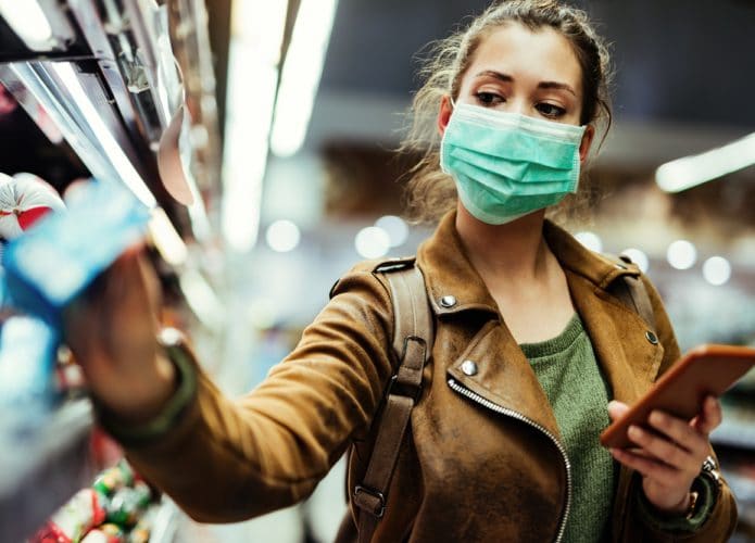 Jovem mulher com máscara facial, usando telefone celular e fazendo compras no supermercado durante a pandemia do vírus.