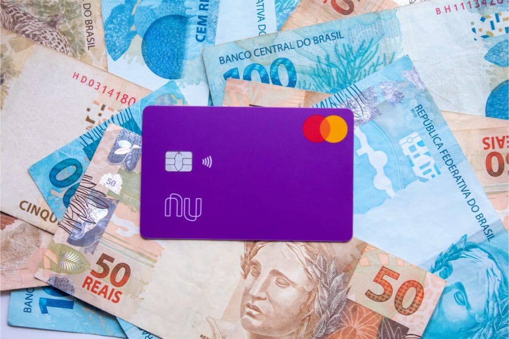 Imagem mostra um fundo com notas de 50 e 100 reais, com o cartão do Nubank à frente. Rumores falam em abertura de capital da empresa