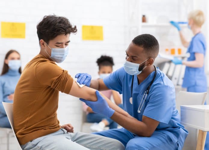 Imagem mostra um enfermeiro aplicando uma vacina em um jovem; ao fundo, aparecem outros enfermeiros