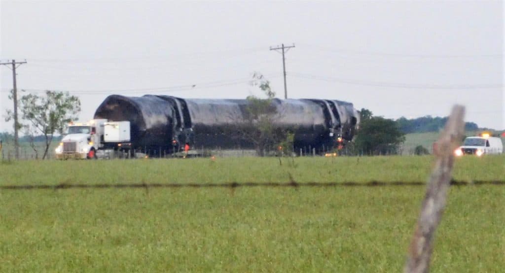 Imagem mostra um propulsor de um foguete Falcon 9, da SpaceX, em trânsito na traseira de um enorme caminhão, seguido de uma van com sirenes acesas