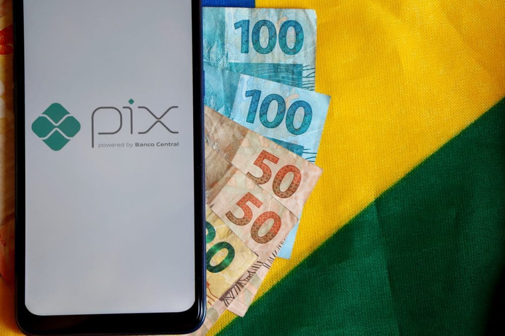 Logo do PIX exibido em smartphone e diversas cédulas sob uma bandeira do Brasil