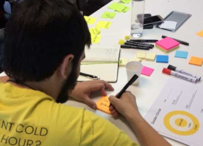 Homem aparece escrevendo em um post-it em cima de uma mesa.