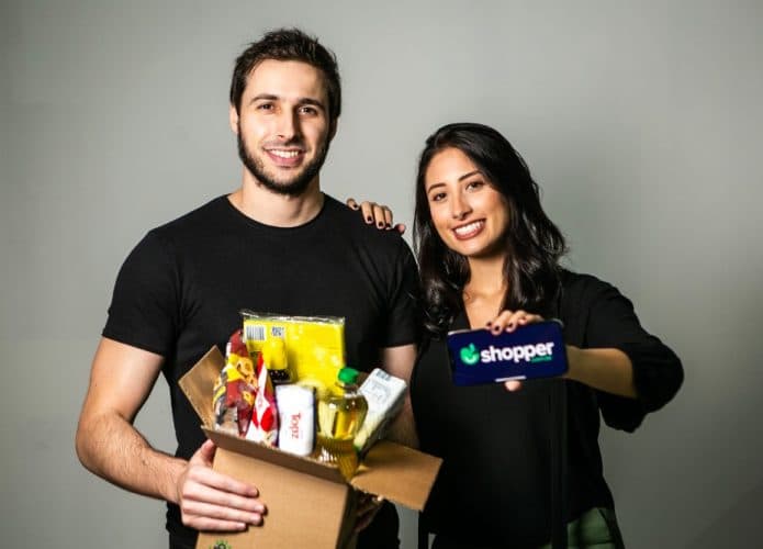 Imagem mostra os cofundadores da startup Shopper, Fábio Rodas e Bruna Vaz; Fábio segura uma caixa com produtos de supermercado, especialidade da empresa, enquanto Bruna segura um smartphone que leva o logo da empresa na tela.