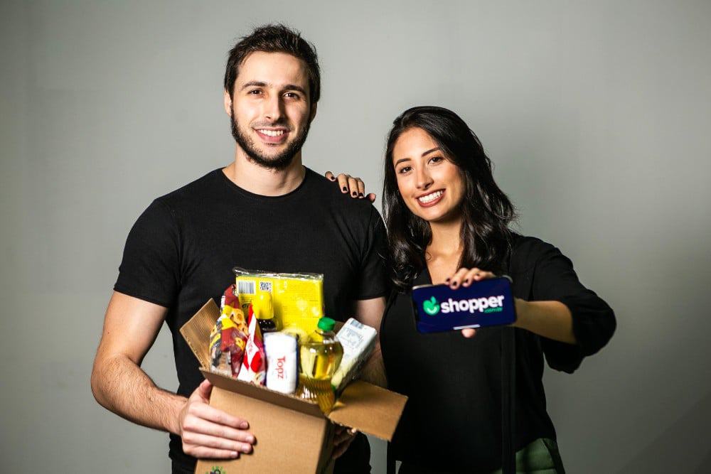 Imagem mostra os cofundadores da startup Shopper, Fábio Rodas e Bruna Vaz; Fábio segura uma caixa com produtos de supermercado, especialidade da empresa, enquanto Bruna segura um smartphone que leva o logo da empresa na tela.