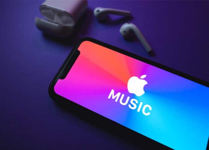 Logomarca do Apple Music é exibida em um smartphone, com dois fones sem fio posicionados ao lado em frente a um fundo azul