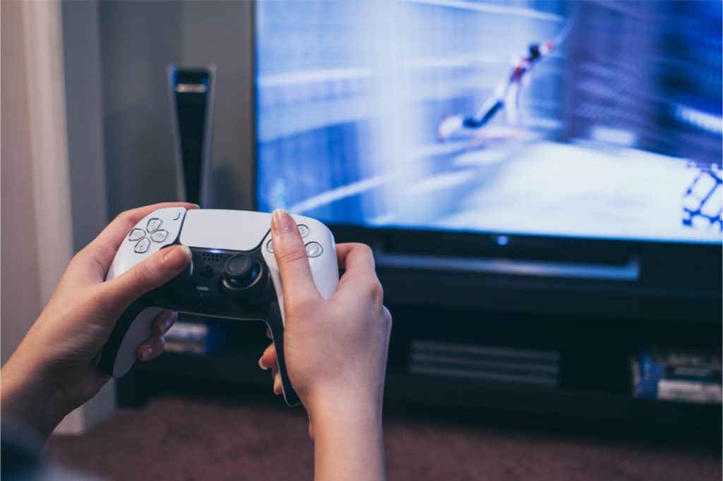 Imagem mostra um controle DualSense do PlayStation 5 segurado por duas mãos, com uma televisão ligada ao fundo, simbolizando uma partida em curso no videogame
