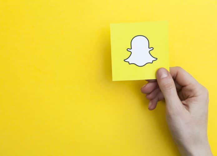 Em fundo amarelo, imagem mostra uma mão segurando um cartão que tem uma ilustração do fantasma, logomarca da rede social Snapchat