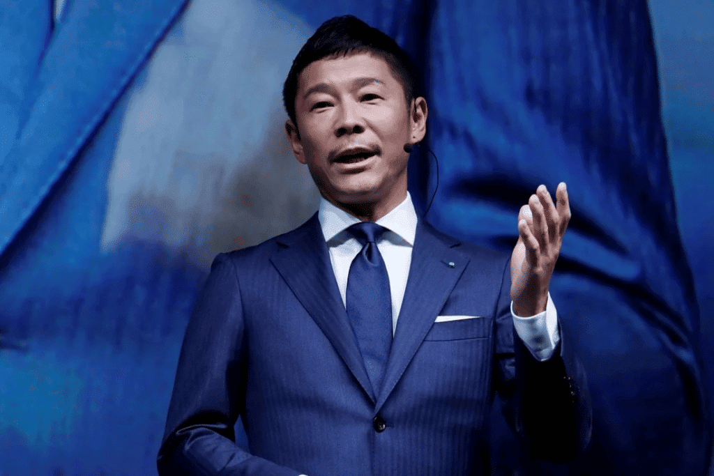 O bilionário japonês Yusaku Maezawa, que viajará para a ISS em dezembro de 2021, é exibido na imagem dando uma palestra, vestindo um paletó azul, com gravata azul escura e portando um microfone de orelha