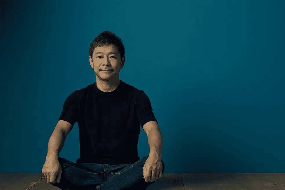 Yusaku Maezawa, bilionário japonês, é mostrado vestindo camiseta preta e calça jeans. Ele está sentado e com as pernas cruzadas, olhando para a câmera e sorrindo. Por trás dele, apenas um fundo azul escuro