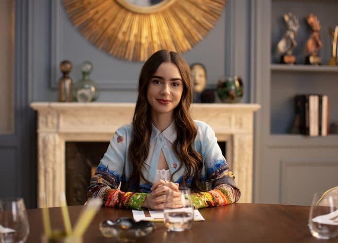 Lily Collins caracterizada como Emily, em cena da série Emily em Paris, da Netflix. A atriz aparece sentada à mesa, olhando para a frente. Atriz deve mudar o visual para viver a boneca Polly Pocket.