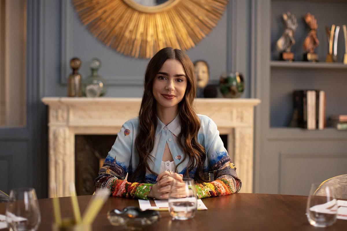 Lily Collins caracterizada como Emily, em cena da série Emily em Paris, da Netflix. A atriz aparece sentada à mesa, olhando para a frente. Atriz deve mudar o visual para viver a boneca Polly Pocket.