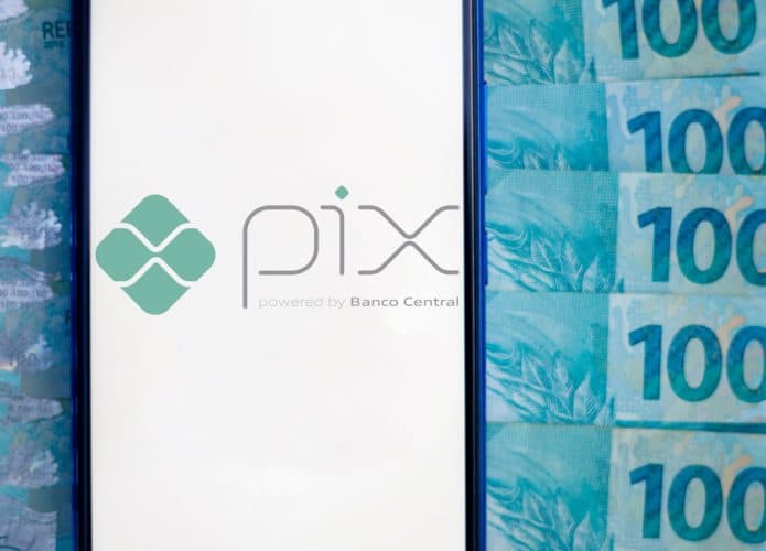 Imagem mostra a tela de um smartphone exibindo o logotipo do sistema de pagamentos instantâneo do Banco Central, o PIX; ao fundo aparecem várias notas de cem reais.