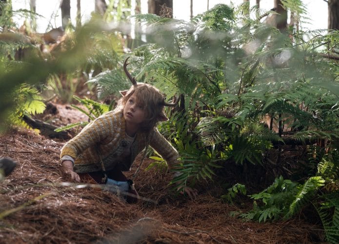 A imagem mostra o ator Christian Convery caracterizado como Gus, uma criança híbrida de humano e cervo, rastejando em uma floresta.